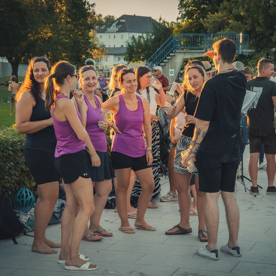 Eine Gruppe von Frauen in gleichen lila Tanktops und schwarzen Shorts steht in einem Park und unterhält sich mit einem Mann, der ein Buch in der Hand hält.