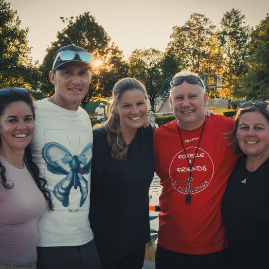 Fünf Personen lächeln und posieren für ein Foto in der Nähe eines Pools, wobei ein Mann ein Hemd mit Schmetterlingsdruck trägt und eine Frau ein rotes Hemd mit 'Forelle & Friends' Text.