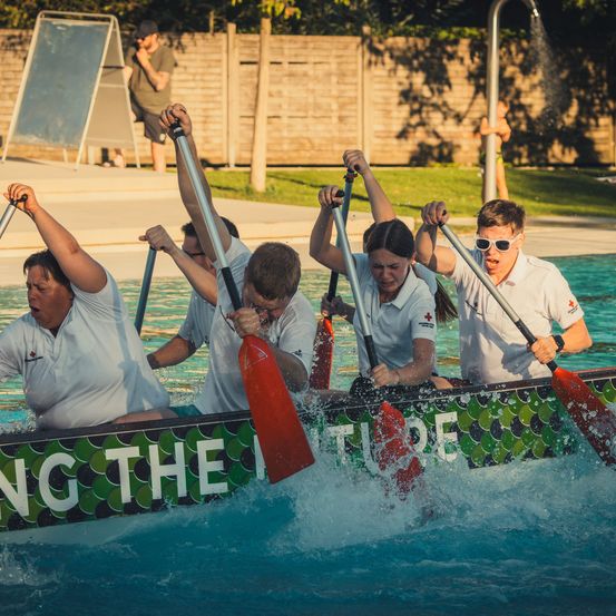 Eine Gruppe von Menschen in weißen Hemden paddelt ein Boot in einem Pool und erzeugt Spritzer. Das Boot ist mit grünen Schuppen und den Worten 'Schwimmt die Zukunft' verziert.