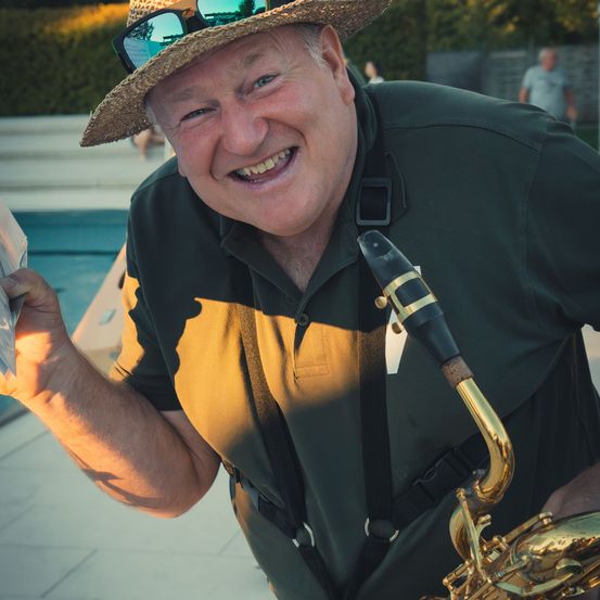 Ein älterer Mann mit Strohhut und Sonnenbrille hält ein Saxophon und lächelt in der Nähe eines Pools.