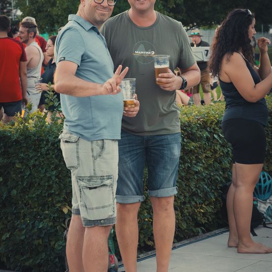 Zwei Männer stehen bei einer Outdoor-Veranstaltung zusammen, einer hält ein Glas Bier und der andere macht ein Friedenszeichen. Eine Frau steht hinter ihnen, teilweise sichtbar.