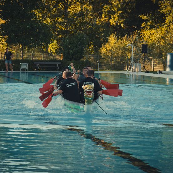 Eine Gruppe von Menschen in einem Drachenboot paddelt in einem Pool mit Bäumen im Hintergrund.