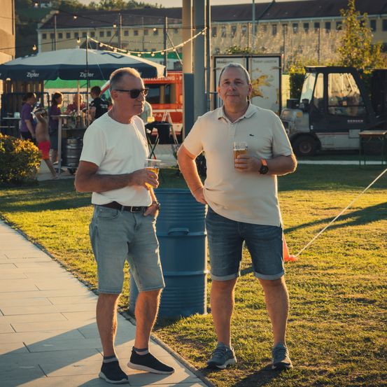 Zwei Männer stehen auf einem Bürgersteig, einer hält ein Glas Bier in der Hand, der andere trägt eine Uhr, vor einem blauen Fass und einem Grasbereich.