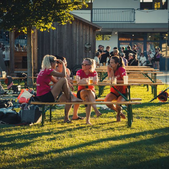 Drei Frauen in passenden roten Shirts sitzen an einem Picknicktisch in einem Park, mit einem Gebäude und Menschen im Hintergrund.