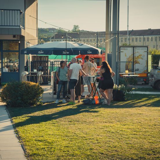 Eine Gruppe von Menschen steht unter einem Schirm in einem Grasbereich, mit einem Food Truck und Gebäuden im Hintergrund.