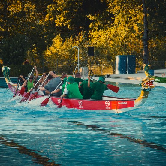 Eine Gruppe von Menschen in grünen Shirts paddelt ein rotes Drachenboot in einem Pool, mit Bäumen und einem Lautsprecher im Hintergrund.