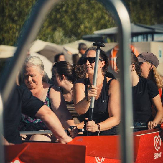 Eine Gruppe von Frauen in einem roten Boot, mit Sonnenbrillen und Paddeln, mit einem unscharfen Hintergrund von Bäumen und Gebäuden.