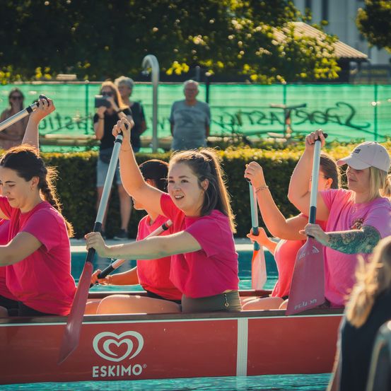 Eine Gruppe von Frauen in pinken Shirts paddelt ein rotes Eskimo-Boot im Pool, während Zuschauer dahinter zusehen.