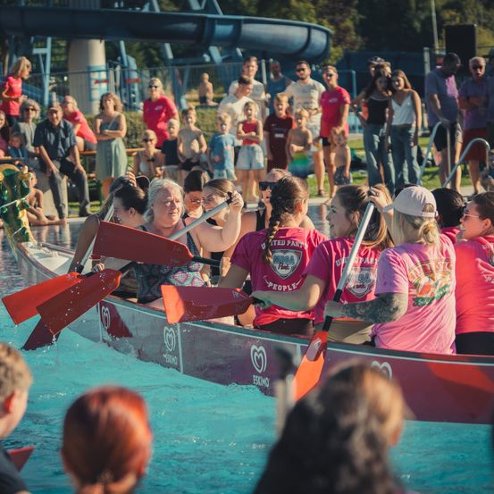 Eine Gruppe von Frauen in rosa Shirts paddelt ein rotes Kanu im Pool, während Zuschauer von der Seite zusehen.