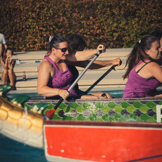 Drei Frauen in lila Tanktops rudern ein Drachenboot im Pool, während Zuschauer von der Seite zusehen.