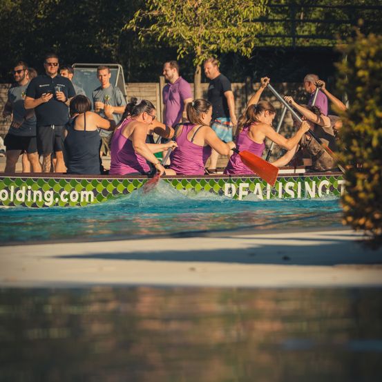Eine Gruppe von Frauen in lila Shirts rudert in einem Pool, während Zuschauer zusehen. Das Boot trägt den Text 'Dealising'.