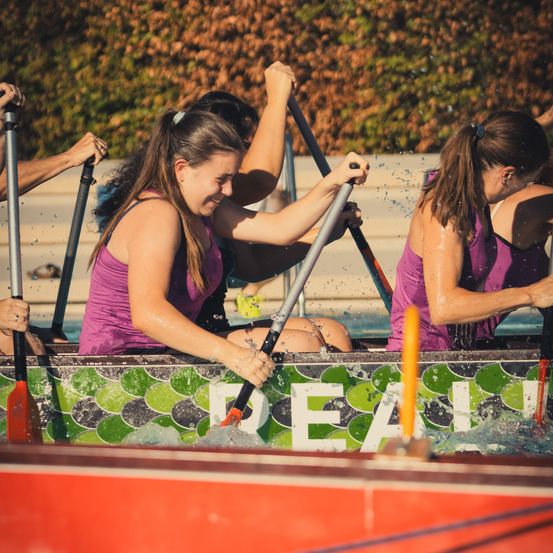 Eine Gruppe von Frauen in lila Tanktops rudert ein Boot und spritzt Wasser. Sie lächeln und scheinen Spaß zu haben.