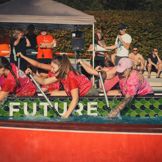 Eine Gruppe von Frauen in roten Shirts paddelt ein Boot mit der Aufschrift 'Future' in einem Pool, während Zuschauer zusehen.