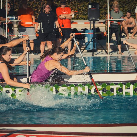 Eine Gruppe von Frauen rudert mit einem Boot im Pool, während Zuschauer von den Seitenlinien zuschauen. Das Boot hat grüne und weiße Designs.