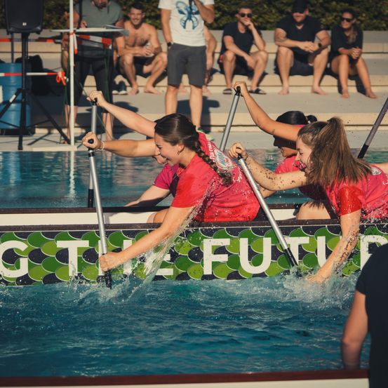 Eine Gruppe von Frauen in roten Shirts paddelt ein Boot im Pool, während Zuschauer von den Rändern zuschauen.