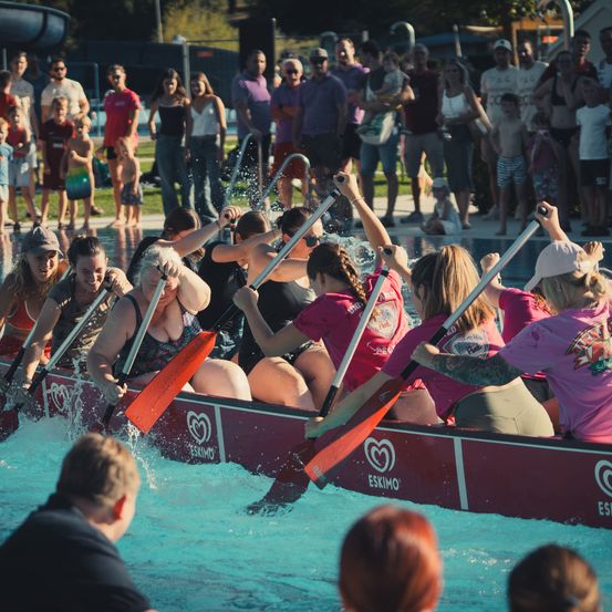 Eine Gruppe von Frauen in Badeanzügen paddelt mit einem roten Boot im Pool, umgeben von einer jubelnden Menge.