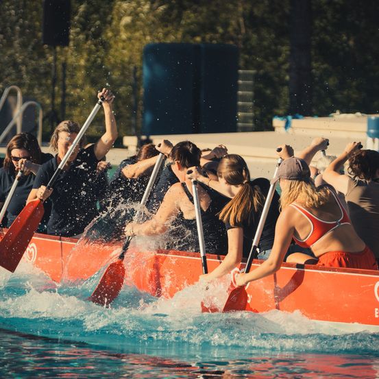 Eine Gruppe von Frauen in einem roten Boot paddelt energisch und verursacht Wasserspritzer um sie herum. Sie tragen alle Sonnenbrillen und einige haben Hüte auf. Das Boot ist mit dem Buchstaben E gekennzeichnet.