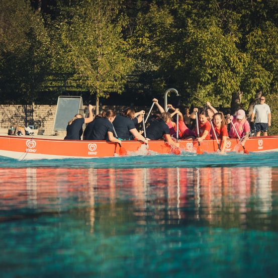Eine Gruppe von Menschen in roten Shirts rudert ein Boot in einem Pool mit Bäumen im Hintergrund.