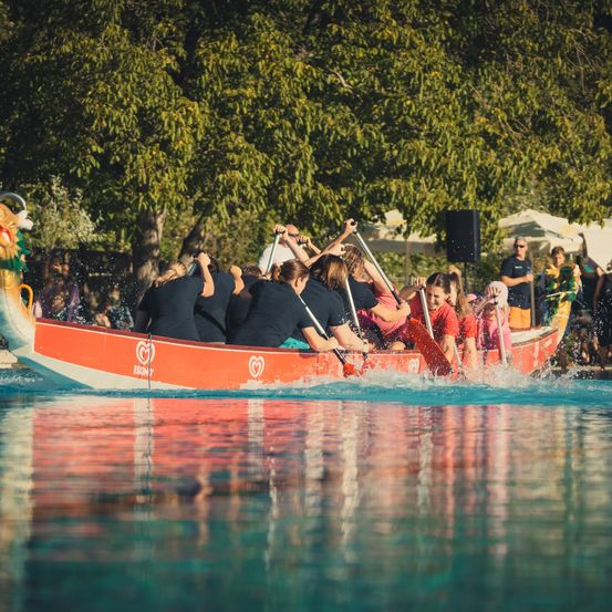 Eine Gruppe von Menschen in einem roten Drachenboot paddelt in einem Pool, während Zuschauer von den Seitenlinien zusehen.