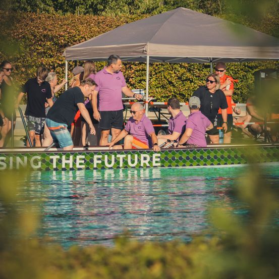 Eine Gruppe von Menschen in lila Shirts steht um ein Boot in einem Pool herum, mit einem Baldachin im Hintergrund.
