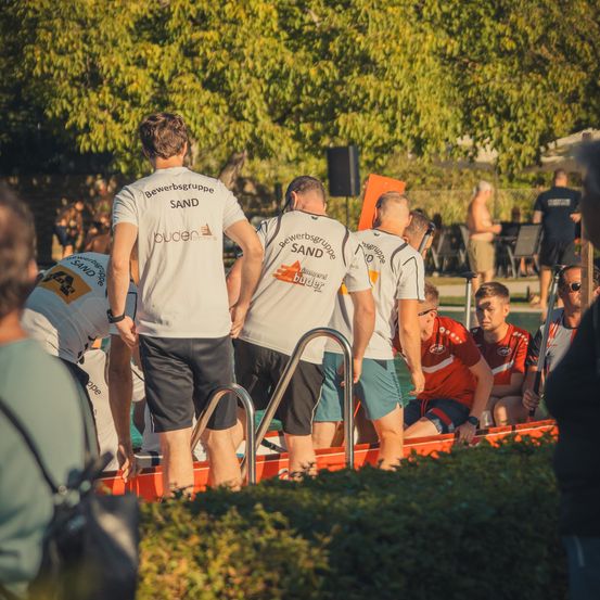 Eine Gruppe von Menschen in weißen T-Shirts mit dem Wort Sand darauf stehen in einem roten Boot. Sie nehmen wahrscheinlich an einer Wassersportveranstaltung teil.