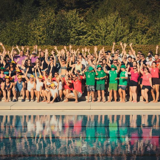 Eine große Gruppe von Menschen in Badekleidung und Hüten, die am Pool stehen und sitzen und ihre Hände in die Luft heben.