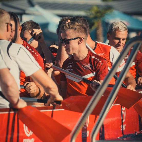 Eine Gruppe von Männern in roten und weißen Trikots rudert ein rotes Boot, wobei einer eine Sonnenbrille trägt und ein anderer sein Haar zurechtrückt.