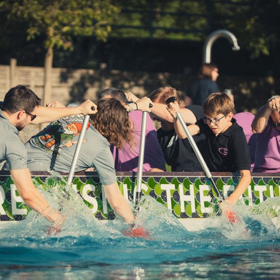 Eine Gruppe von Menschen rudert in einem Pool, wobei Wasserspritzer entstehen. Sie tragen Freizeitkleidung und halten Paddel.