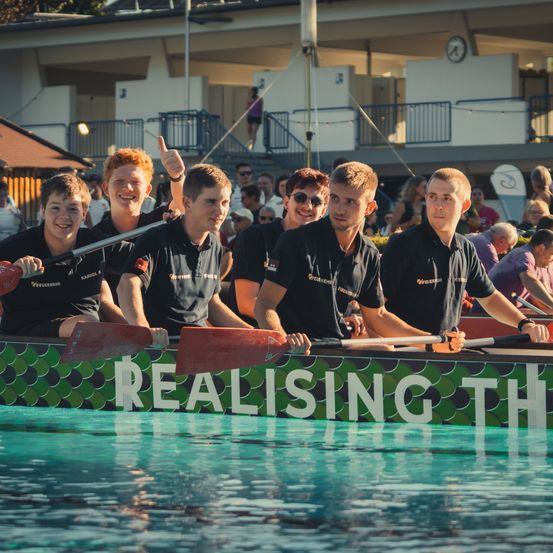 Eine Gruppe junger Männer in schwarzen Shirts paddelt ein Drachenboot im Pool, mit 'Realising the' auf der Seite. Dahinter schauen Zuschauer zu.