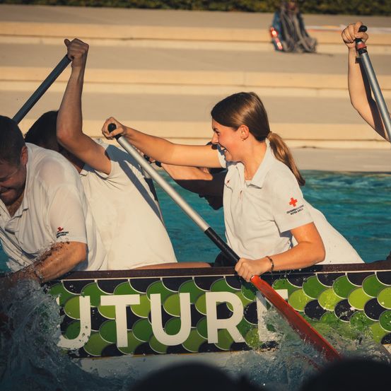 Eine Gruppe von Menschen in weißen Hemden rudert ein Boot mit dem Wort 'TUR' im Pool. Sie lächeln und scheinen Spaß zu haben.