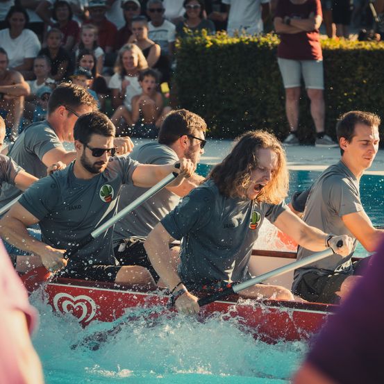 Eine Gruppe von Männern rudert mit einem roten Boot im Pool, während eine Menge zuschaut. Die Männer tragen graue Shirts und schwarze Shorts. Das Boot hat ein rotes Herzdesign.