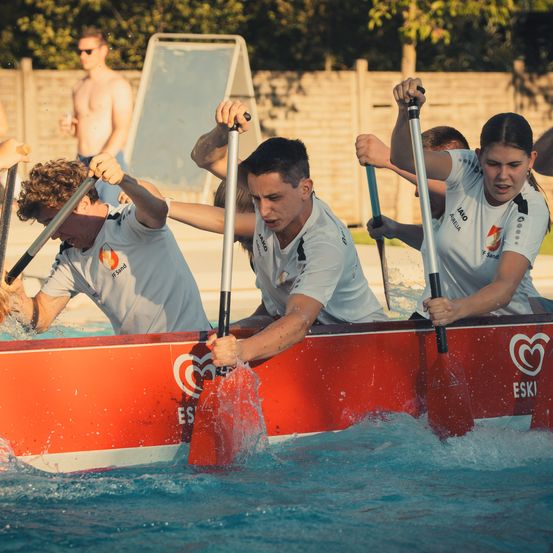 Eine Gruppe von Menschen rudert in einem Pool in einem roten Boot und trägt weiße T-Shirts mit Logos. Das Wasser spritzt um sie herum.