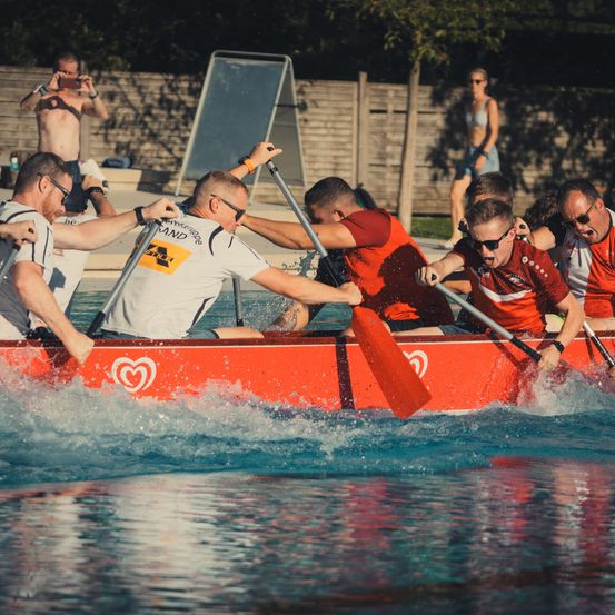 Eine Gruppe von Männern in roten und weißen Shirts paddelt ein rotes Boot in einem Pool, mit Wasserspritzern und einem Herzlogo auf dem Boot.