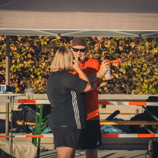 Ein Mann und eine Frau stehen unter einem Zelt, der Mann hält eine Hupe, während die Frau hineinbläst. Sie sind in Sportkleidung gekleidet.