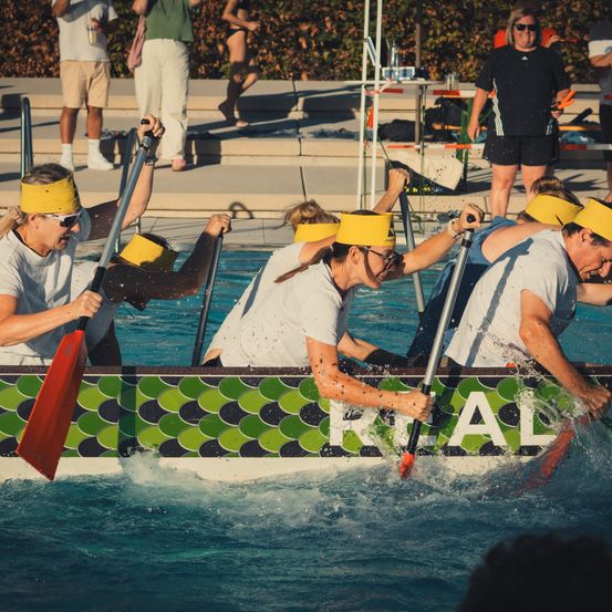 Eine Gruppe von Menschen mit gelben Stirnbändern rudert in einem Pool. Sie halten Paddel und das Boot hat grüne und schwarze Designs.