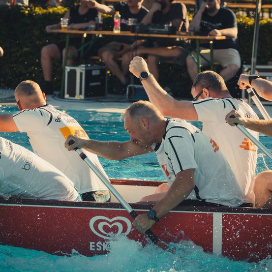 Eine Gruppe von Männern in weißen Shirts rudert mit einem roten Boot im Pool, wobei Wasserspritzer um sie herum sind. Zuschauer sitzen an Tischen im Hintergrund.