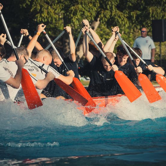 Eine Gruppe von Menschen in schwarzen Shirts rudert mit orangefarbenen Paddeln in einem Boot auf dem Wasser.