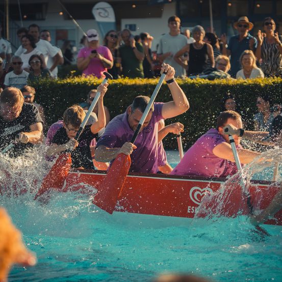 Eine Gruppe von Menschen paddelt in einem roten Boot im Pool, während Zuschauer von der Seite zusehen.