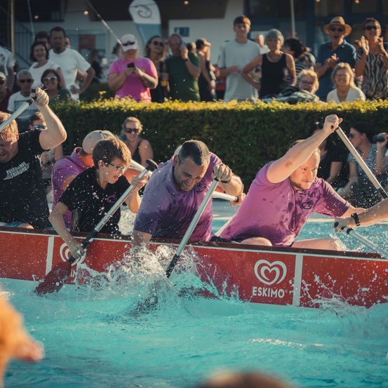 Eine Gruppe von Männern in einem roten Boot mit Paddeln, die in einem Pool Rennen fahren, während eine Menge zuschaut.
