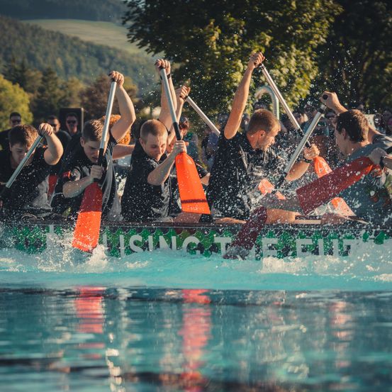 Eine Gruppe von Männern in schwarzen Shirts paddelt ein Boot in einem Pool und erzeugt Wasserspritzer. Das Boot hat ein grünes und weißes Design.