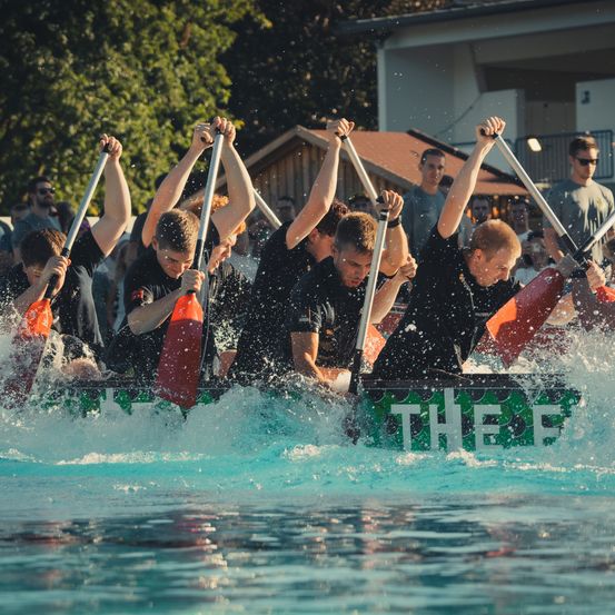 Eine Gruppe von Männern in schwarzen T-Shirts paddelt ein mit 'The F' beschriftetes Boot in einem Pool und erzeugt Spritzer. Zuschauer beobachten von der Seite.