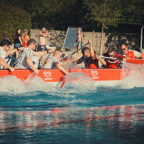 Eine Gruppe von Menschen rudert ein rotes Boot in einem Pool und erzeugt Spritzer. Zuschauer beobachten von der Seite.