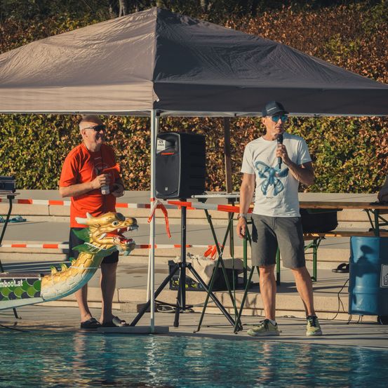 Zwei Männer stehen unter einem Zelt am Pool, einer hält ein Mikrofon und der andere eine Flasche. Eine Drachenstatue steht am Poolrand.