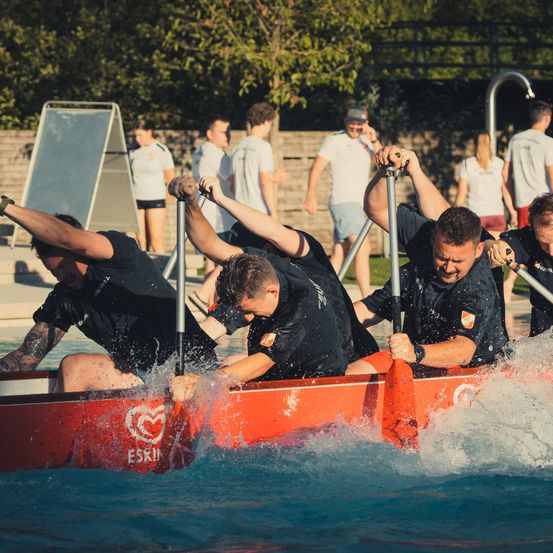 Eine Gruppe von Männern in schwarzen Shirts rudert ein rotes Boot in einem Pool, wobei Wasser um sie herum spritzt.