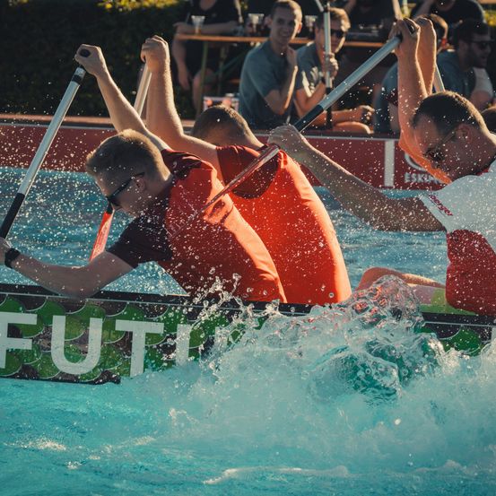 Vier Männer in roten Rettungswesten paddeln ein Drachenboot im Pool und verursachen Spritzer. Zuschauer beobachten von der Seite.