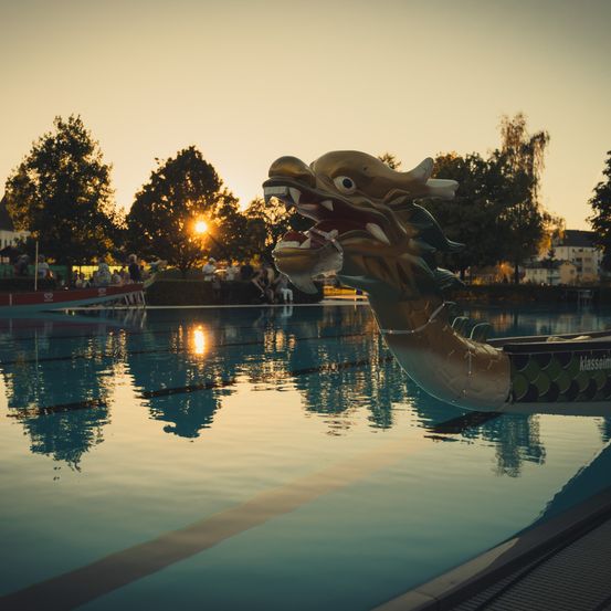 Ein drachenförmiges Boot ist in einem Pool verankert, mit einem Sonnenuntergang im Hintergrund. Bäume und Gebäude sind in der Ferne zu sehen.