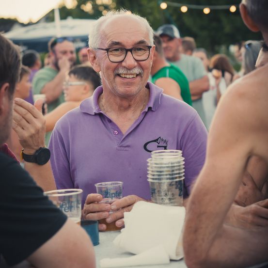 Ein älterer Mann mit Brille und einem lila Poloshirt lächelt und hält einen Becher bei einer überfüllten Outdoor-Veranstaltung.