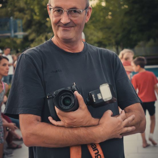 Ein Mann mit Brille und schwarzem T-Shirt hält eine Kamera mit Blitz vor einem verschwommenen Hintergrund mit Menschen.