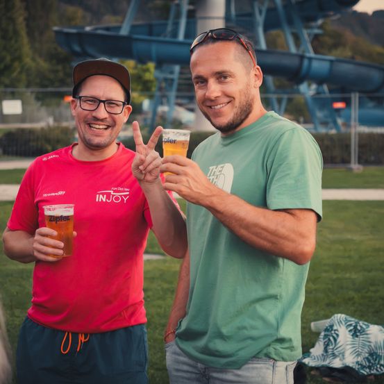 Zwei Männer stehen in einem Park, einer in einem roten Shirt und der andere in einem grünen Shirt, beide halten Biergläser und lächeln. Hinter ihnen ist eine Wasserrutsche und ein Zaun.