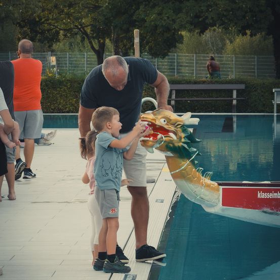 Ein älterer Mann und zwei Kinder stehen am Pool, der Mann füttert einen drachenförmigen Brunnen mit einer Münze.